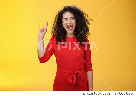 Rebellious meloman leaving in tender girl. Thrilled and carefree curly-haired woman in red dress sticking out tongue and looking right with smile as showing rock-n-roll gesture, enjoying cool music Rebellious meloman leaving in tender girl. Thrilled and carefree curly-haired woman in red dress sticking out tongue and looking right with smile as showing rock-n-roll gesture, enjoying cool music 84663650