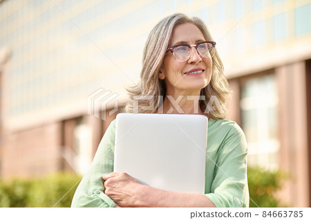 Thoughtful woman in glasses hugging laptop on street Thoughtful woman in glasses hugging laptop on street 84663785