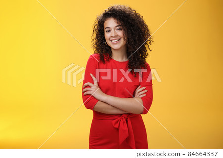 Confident charismatic young curly-haired woman in casual red dress holding hands crossed over chest in self-assured satisfied expression, smiling delighted posing over yellow background 84664337