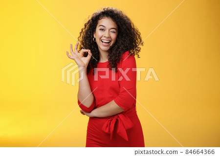 Girl feeling awesome. Friendly-looking excited and upbeat charming girlfriend with curly hair in dress winking playfully and smiling as showing okay gesture having deal and things under control Girl feeling awesome. Friendly-looking excited and upbeat charming girlfriend with curly hair in dress winking playfully and smiling as showing okay gesture having deal and things under control 84664366