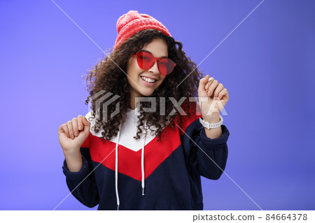 Stylish and feminine girlfriend dancing having fun feeling upbeat as liking weather feeling warm and cozy in stylish red beanie and sunglasses dancing with raised hands and broad smile over blue wall Stylish and feminine girlfriend dancing having fun feeling upbeat as liking weather feeling warm and cozy in stylish red beanie and sunglasses dancing with raised hands and broad smile over blue wall 84664378