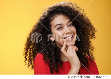 Headshot of cute silly and tender feminine romantic woman with curly hairstyle touching lip with index finger making eyes at camera and smiling as using seduction skills over yellow background Headshot of cute silly and tender feminine romantic woman with curly hairstyle touching lip with index finger making eyes at camera and smiling as using seduction skills over yellow background 84664380