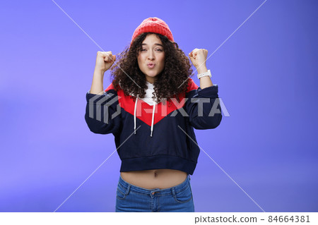 Charming young 20s girl with curly hair in warm beanie raising hands as belly showing under sweatshirt triumphing, celebrating awesome win folding lips from joy and excitement over blue background Charming young 20s girl with curly hair in warm beanie raising hands as belly showing under sweatshirt triumphing, celebrating awesome win folding lips from joy and excitement over blue background 84664381