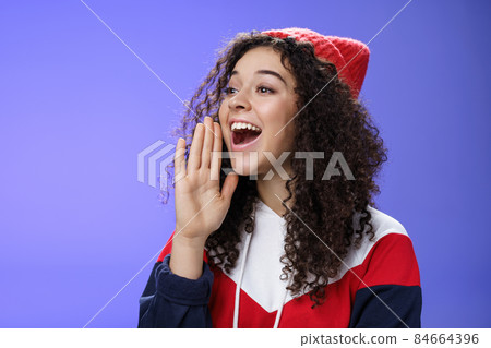 Friend shouting through street to turn attention. Portrait of carefree happy cute woman in beanie and sweatshirt holding palm near opened mouth looking with smile left as calling mate with loud yell Friend shouting through street to turn attention. Portrait of carefree happy cute woman in beanie and sweatshirt holding palm near opened mouth looking with smile left as calling mate with loud yell 84664396