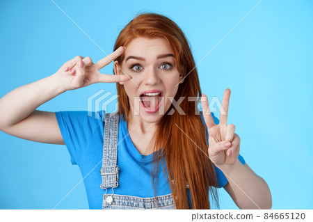 Cute excited redhead enthusiastic girl smiling, look joyful show peace, victory signs near face, optimistic lucky attitude, believe win, stand blue background positive, having fun enjoy vacation 84665120