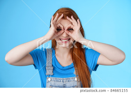 Playful cheerful redhead european woman, long nature red hairstyle, show okay finger glasses look through ok signs, smiling broadly funny grimace, stand blue background carefree enjoy summer 84665155