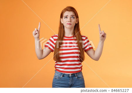 Hesitant upset cute redhead woman weighing choices look doubtful uncertain, frowning thoughtful, ponder decision, look pointing up unsure, have doubts, stand insecure orange background 84665227