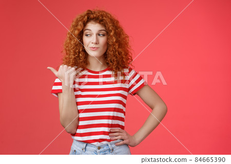 Look that stuff. Redhead curious sassy good-looking curly-haired girl summer striped t-shirt smirking gazing pointing left indicating thumb intriguing product standing red background 84665390
