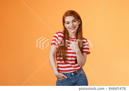 Tender cute feminine redhead lovely girl, tilt head cheerfully smiling, pointing left, showing way, give advice what purchase, advertising good product, introduce shop, orange background 84665476