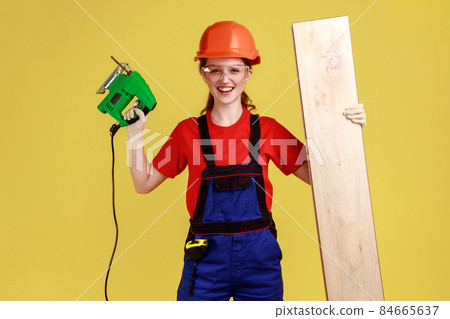 Portrait of young happy positive carpenter woman working with fretsaw, holding wooden panel, wearing overalls and protective helmet. Indoor studio shot isolated on yellow background. Portrait of young happy positive carpenter woman working with fretsaw, holding wooden panel, wearing overalls and protective helmet. Indoor studio shot isolated on yellow background. 84665637