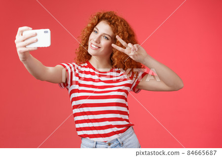 Stylish carefree good-looking redhead curly woman enjoy summer holidays show victory peace gesture eye tilt head cute record video hold smartphone taking selfie red background 84665687