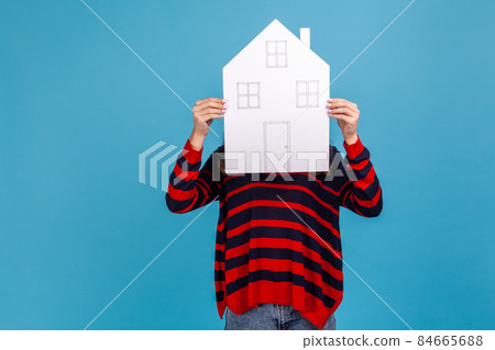 Unknown woman wearing striped casual style sweater, hiding her face behind paper house with drawn windows and door, government housing program. Indoor studio shot isolated on blue background. 84665688
