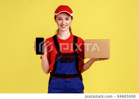 Optimistic delivery woman standing with carton box, parcel for client, showing cell phone with blank screen for promotion, wearing overalls and cap. Indoor studio shot isolated on yellow background. 84665690