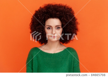 Portrait of serious strict woman with Afro hairstyle wearing green casual style sweater looking at camera with assertive facial expression. Indoor studio shot isolated on orange background. 84665708