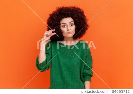 Portrait of woman with Afro hairstyle wearing green casual style sweater showing small size gesture with fingers, looking at camera with disappointment. Indoor studio shot isolated on orange Portrait of woman with Afro hairstyle wearing green casual style sweater showing small size gesture with fingers, looking at camera with disappointment. Indoor studio shot isolated on orange 84665711