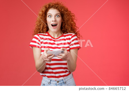 Amused impressed good-looking curly-haired redhead female student unexpectedly win smartphone game look camera astonished gasping open mouth stare camera hold phone horizontal playing app Amused impressed good-looking curly-haired redhead female student unexpectedly win smartphone game look camera astonished gasping open mouth stare camera hold phone horizontal playing app 84665712