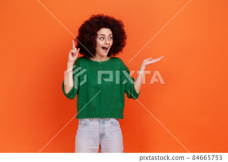 Positive woman with Afro hairstyle wearing green casual style sweater pointing finger up, has idea, presenting area for advertisement. Indoor studio shot isolated on orange background. 84665753
