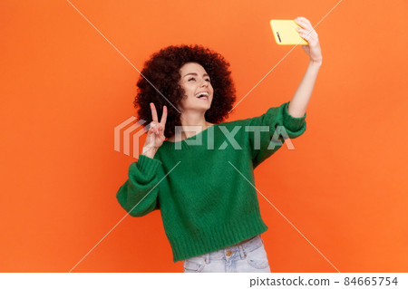 Happy woman with Afro hairstyle wearing green casual style sweater having online conversation with followers in social networks, showing v sign. Indoor studio shot isolated on orange background. 84665754