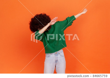 Woman with Afro hairstyle wearing green sweater showing dab dance, popular internet meme pose, celebrating success victory, dabbing trends concept. Indoor studio shot isolated on orange background. 84665759