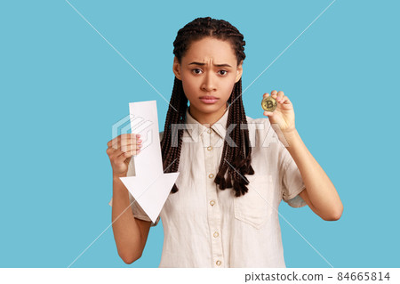 Portrait of dissatisfied woman with dreadlocks showing gold bitcoin and arrow pointing down, downgrade of electronic currency, wearing white shirt. Indoor studio shot isolated on blue background. 84665814