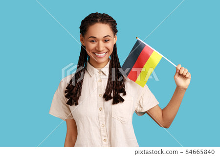 Portrait of pleasant looking woman with black dreadlocks holding Germany flag, celebrating Day of Germany - 3th October, wearing white shirt. Indoor studio shot isolated on blue background. Portrait of pleasant looking woman with black dreadlocks holding Germany flag, celebrating Day of Germany - 3th October, wearing white shirt. Indoor studio shot isolated on blue background. 84665840