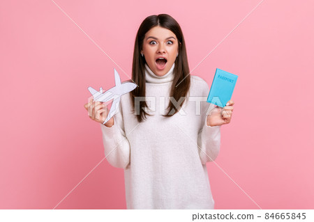 Shocked astonished young woman holding passport and paper plane in hands planning vacation abroad, wearing white casual style sweater. Indoor studio shot isolated on pink background. Shocked astonished young woman holding passport and paper plane in hands planning vacation abroad, wearing white casual style sweater. Indoor studio shot isolated on pink background. 84665845