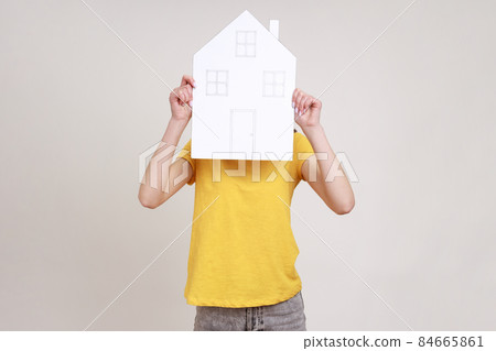 Unknown female in yellow T-shirt hiding her face behind paper house with drawn windows and door, government housing program, dreaming of new house. Indoor studio shot isolated on gray background. 84665861