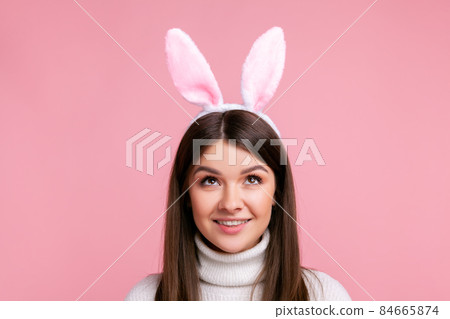 Young female with bunny ears looking up with playful attractive smile, childish carefree expression, wearing white casual style sweater. Indoor studio shot isolated on pink background. Young female with bunny ears looking up with playful attractive smile, childish carefree expression, wearing white casual style sweater. Indoor studio shot isolated on pink background. 84665874