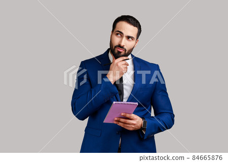 Young adult businessman standing with thoughtful expression, holding paper notebook, pondering business idea, future plans, wearing official style suit. Indoor studio shot isolated on gray background. 84665876