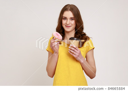 Sweet temptation. Portrait of happy pretty teenager girl holding appetizing donut and take away coffee, high-calorie junk food for breakfast. Indoor studio shot isolated on gray background. Sweet temptation. Portrait of happy pretty teenager girl holding appetizing donut and take away coffee, high-calorie junk food for breakfast. Indoor studio shot isolated on gray background. 84665884