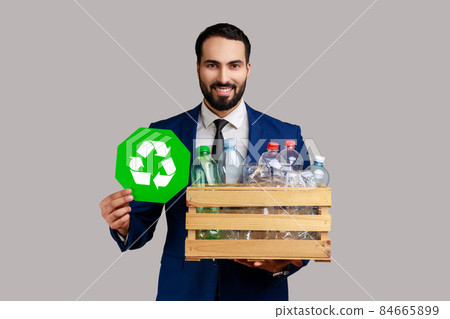 Bearded man holding box with plastic bottles and recycling green symbol, sorting his rubbish, saving ecology, wearing official style suit. Indoor studio shot isolated on gray background. Bearded man holding box with plastic bottles and recycling green symbol, sorting his rubbish, saving ecology, wearing official style suit. Indoor studio shot isolated on gray background. 84665899