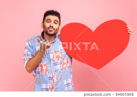 Portrait of happy romantic bearded man with big red heart in hands, looking at camera, sending air kisses, wearing blue casual style shirt. Indoor studio shot isolated on pink background. Portrait of happy romantic bearded man with big red heart in hands, looking at camera, sending air kisses, wearing blue casual style shirt. Indoor studio shot isolated on pink background. 84665908