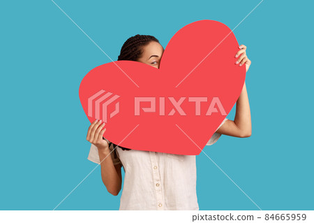 Portrait of unknown woman holding big red heart in hands, peeping from love symbol, being shy to tell about her feelings, wearing white shirt. Indoor studio shot isolated on blue background. 84665959
