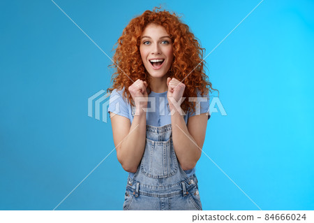 Cheerful optimistic lucky redhead curly-haired attractive woman cheering clench fists joyfully smiling broadly cheering watching game supportive encourage keep up motivated blue background 84666024