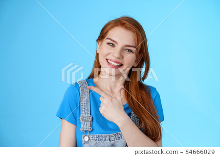 Friendly cheerful caucasian red woman, tilt head smiling pleasantly, suggest good copy space, pointing left introduce promo, give advice, indicating awesome link, pointing blue background 84666029