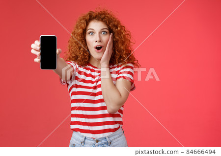 Amused impressed redhead good-looking curly-haired excited girl share awesome social media page show smartphone screen amazed touch cheek thrilled affected awesome app red background 84666494