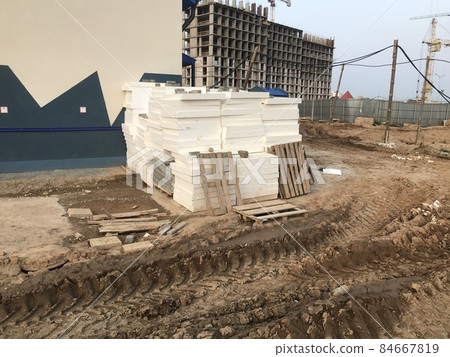 dirt at a construction site. sand furrows, tracks from heavy vehicles. next to it are building materials, polystyrene, wooden pallets. against the background of houses under construction, high cranes 84667819