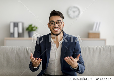 Portrait of young middle eastern psychologist gesturing and smiling at camera, sitting on comfy couch in office Portrait of young middle eastern psychologist gesturing and smiling at camera, sitting on comfy couch in office 84668327