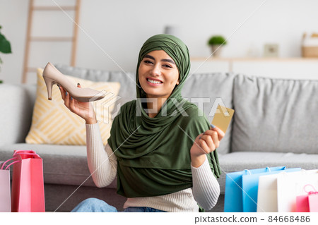 Young Arab woman in hijab showing credit card and shoe, sitting on floor among gift bags, buying footwear on internet Young Arab woman in hijab showing credit card and shoe, sitting on floor among gift bags, buying footwear on internet 84668486