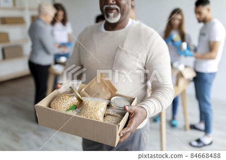 Humanitarian aid. African american elderly man holding box with donations food, volunteers packing boxes with products Humanitarian aid. African american elderly man holding box with donations food, volunteers packing boxes with products 84668488