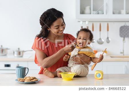 Childcare. Loving Black Mom Feeding Her Baby Son From Spoon In Kitchen Childcare. Loving Black Mom Feeding Her Baby Son From Spoon In Kitchen 84668522