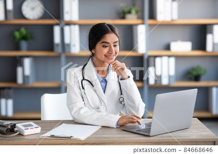 Smiling young indian female doctor in white coat works at laptop at workplace in clinic office interior Smiling young indian female doctor in white coat works at laptop at workplace in clinic office interior 84668646