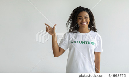 Look there. Happy black female volunteer pointing finger aside and smiling, showing free space over light wall, panorama 84668753