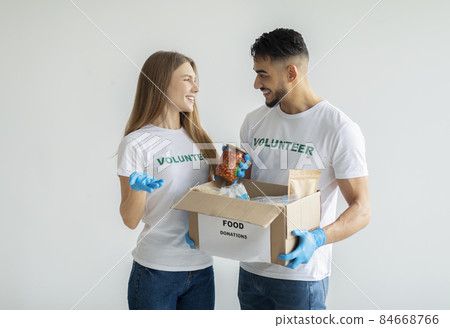 Charity donation center. Young diverse volunteers holding box with products, sorting humatiranian aid over light wall 84668766