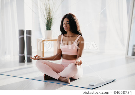 Peaceful young black woman sitting in lotus pose, meditating, reaching inner peace and harmony on mat at home Peaceful young black woman sitting in lotus pose, meditating, reaching inner peace and harmony on mat at home 84668784
