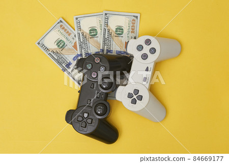 Game console controller and pile of money. Gamepad and us dollars. Wireless game pad on dollar bills. Concept of game addiction, spending money on online games, earning money on gaming streaming 84669177