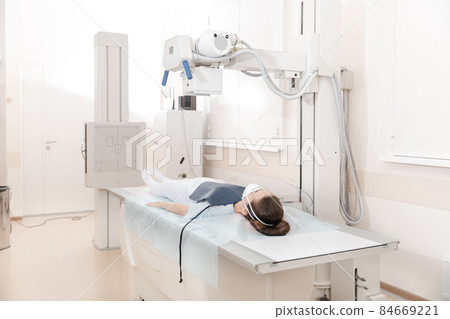 Doctor taking x-ray of patient lying on gurney. Hospital radiology room. Technician adjusting an x-ray machine. Female lying on a bed is scanning chest, heart, lungs in modern clinic office. Doctor taking x-ray of patient lying on gurney. Hospital radiology room. Technician adjusting an x-ray machine. Female lying on a bed is scanning chest, heart, lungs in modern clinic office. 84669221