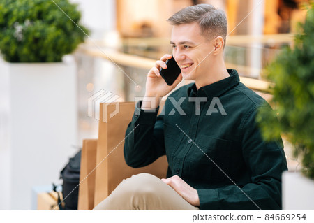 Portrait of handsome smiling young man talking on mobile phone sitting on bench in shopping mall looking away. Portrait of handsome smiling young man talking on mobile phone sitting on bench in shopping mall looking away. 84669254