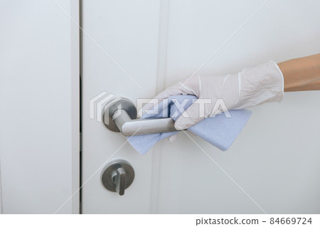 Cleaning door handle with blue wipe in white gloves. Sanitize surfaces prevention in hospital and public spaces against corona virus. Woman hand using towel for cleaning home room door link. 84669724