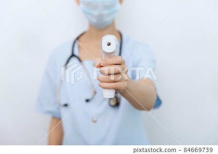 Temperature measurement gun in doctor hands. Close-up shot of doctor wearing protective surgical mask ready to use infrared isometric thermometer gun to check body temperature for virus symptoms. Temperature measurement gun in doctor hands. Close-up shot of doctor wearing protective surgical mask ready to use infrared isometric thermometer gun to check body temperature for virus symptoms. 84669739